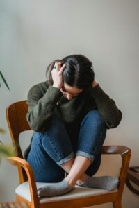 A young woman sitting on a chair indoors, holding her head in distress.
